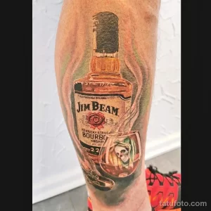 Фото тату на ноге с реалистичным изображением бутылки Jim Beam и черепа в стакане. Детализированный рисунок и коричнево-красная цветовая гамма.