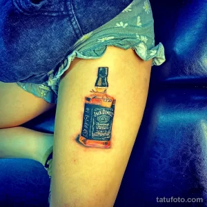 Фото тату на бедре с реалистичным изображением бутылки Jack Daniel's. Детализированный рисунок и коричнево-черная цветовая гамма.