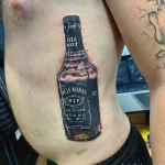 Фото тату на груди с изображением бутылки виски Old No.7 и надписью "Uncle Kieran" с датами рождения и смерти. Памятный рисунок.