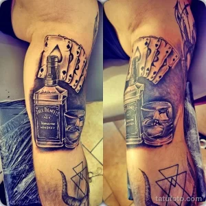 Фото тату на руке с изображением бутылки виски Jack Daniel's, игральных карт, стакана виски и черепов. Реалистичный стиль, темные тона.