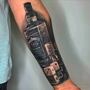 Фото тату на руке: реалистичное изображение бутылки виски Jack Daniel's с игральными костями, выполненное в черно-серых тонах.