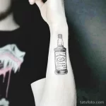 Черно-белая татуировка бутылки виски Jack Daniel's, выполненная на предплечье. Лаконичный рисунок татуировки в стиле графика.
