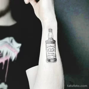 Черно-белая татуировка бутылки виски Jack Daniel's, выполненная на предплечье. Лаконичный рисунок татуировки в стиле графика.