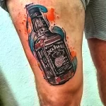 Фотография цветной татуировки на ноге с изображением бутылки Jack Daniel's и брызг краски. Реалистичный рисунок татуировки.