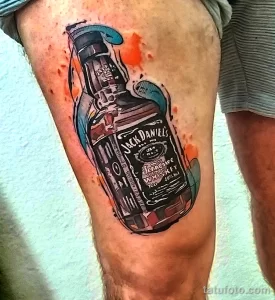 Фотография цветной татуировки на ноге с изображением бутылки Jack Daniel's и брызг краски. Реалистичный рисунок татуировки.