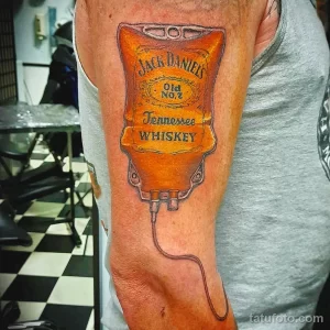 Фотография цветной татуировки на руке с изображением бутылки виски Jack Daniel's и крана для розлива. Детализированный рисунок татуировки.