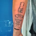 Фото тату на руке: бутылка Jack Daniel's, выполненная в стиле эскиза. Черно-белый рисунок с неровными линиями и эффектом незаконченности.