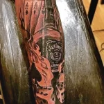 Фото тату на руке: бутылка Jack Daniel's в огне с игральными костями. Детализированный рисунок в черно-серых тонах с яркими акцентами.