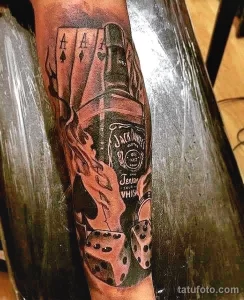 Фото тату на руке: бутылка Jack Daniel's в огне с игральными костями. Детализированный рисунок в черно-серых тонах с яркими акцентами.