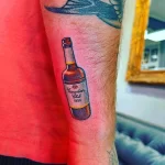 Фото тату минималистичного рисунка бутылки Canadian Club на руке, выполненного в традиционном стиле с использованием ярких цветов и четких линий.