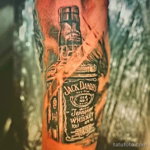 Фото тату на руке: реалистичное изображение бутылки виски Jack Daniel's. Детализированный черно-белый рисунок татуировки.