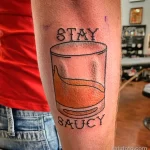 Фото тату на руке: стакан с оранжевым напитком и надпись "Stay Saucy". Простой, минималистичный рисунок татуировки.