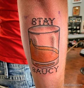 Фото тату на руке: стакан с оранжевым напитком и надпись "Stay Saucy". Простой, минималистичный рисунок татуировки.