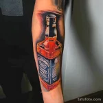 Фото тату на предплечье: реалистичное изображение бутылки виски Jack Daniel's, выполненное в ярких цветах. Детализированный рисунок татуировки.