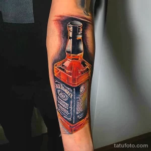 Фото тату на предплечье: реалистичное изображение бутылки виски Jack Daniel's, выполненное в ярких цветах. Детализированный рисунок татуировки.