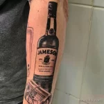 Фото тату реалистичной бутылки виски Jameson с банкнотами. Детализированный рисунок татуировки на руке.