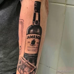 Фото тату реалистичной бутылки виски Jameson с банкнотами. Детализированный рисунок татуировки на руке.