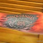 Фото тату Jack Daniel's на руке. Изображена этикетка виски Jack Daniel's в стиле олдскул, с использованием ярких цветов и четких контуров.