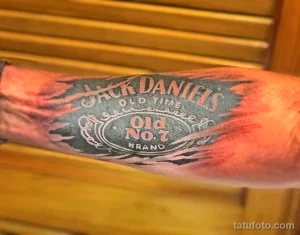 Фото тату Jack Daniel's на руке. Изображена этикетка виски Jack Daniel's в стиле олдскул, с использованием ярких цветов и четких контуров.
