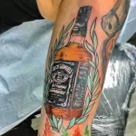 Фото тату Jack Daniel's на руке. Изображена бутылка виски с этикеткой, окруженная зелеными листьями, выполненная в ярких цветах.