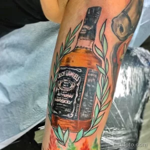 Фото тату Jack Daniel's на руке. Изображена бутылка виски с этикеткой, окруженная зелеными листьями, выполненная в ярких цветах.