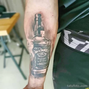 Фото тату Jack Daniel's на предплечье. Черно-белый рисунок бутылки виски с этикеткой, детально проработанный и реалистичный.