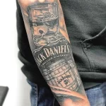 Фото татуировки на руке с изображением этикетки Jack Daniel's, бочки виски и черепа, выполненное в черно-серых тонах.