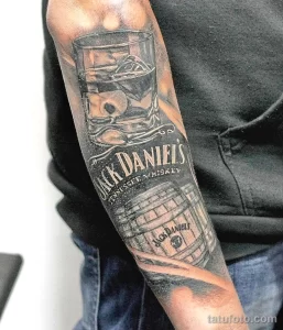 Фото татуировки на руке с изображением этикетки Jack Daniel's, бочки виски и черепа, выполненное в черно-серых тонах.