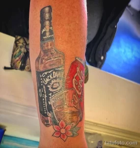 Фото тату на ноге с реалистичным изображением бутылок Jack Daniel's и Coca-Cola, украшенных розовым цветком.