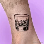 Фото тату на ноге: минималистичный рисунок стакана виски с кубиками льда, выполненный в технике line work.