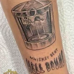 Фото тату на предплечье: рисунок стакана виски с надписью "Whiskey Bent Hell Bound", выполненный в графическом стиле.