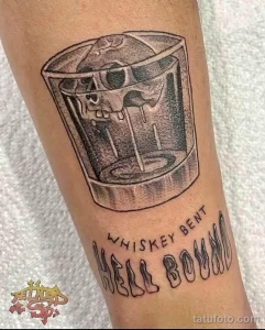 Фото тату на предплечье: рисунок стакана виски с надписью "Whiskey Bent Hell Bound", выполненный в графическом стиле.