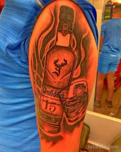 Фото тату на плече: реалистичное изображение бутылки виски Glenfiddich 15 с логотипом и стаканом.