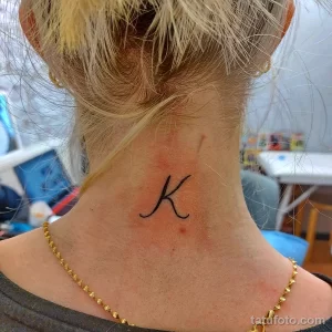 Фото минималистичной татуировки на затылке: маленькая буква 'K', выполненная тонкими линиями черного цвета. Простой и элегантный дизайн.