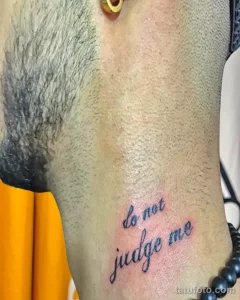 Фото тату: надпись "Do not judge me" на боку, выполненная каллиграфическим шрифтом в синем цвете.