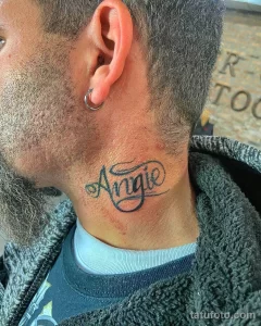 Фото тату с именем 'Angie', выполненным изящным каллиграфическим шрифтом на шее мужчины. Черный рисунок татуировки выглядит стильно и минималистично.