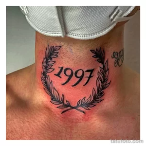 Фото тату на шее: минималистичная татуировка с датой рождения "1997", заключенной в лавровый венок, выполненная в черном цвете.