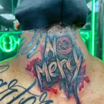Фото тату с надписью 'No Mercy' на затылке, выполненное в ярких красных и черных тонах с брызгами краски. Динамичный рисунок татуировки.