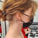 Фотография минималистичной татуировки креста на шее девушки. Татуировка выполнена черными чернилами и имеет простой, лаконичный дизайн.