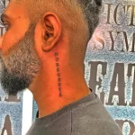Фотография татуировки надписи "No Regrets" на шее мужчины с бородой. Надпись выполнена черными чернилами и расположена вертикально вдоль шеи.