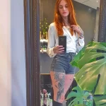 Фото девушки с татуировками на руках и ногах, демонстрирующей свои тату в зеркале, в интерьере тату-салона.