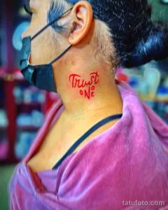 Фото тату: Надпись "Trust One" красными чернилами на шее мужчины. Простая и лаконичная татуировка в стиле леттеринг.