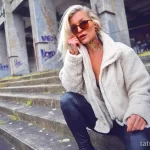 Фото тату: женщина в стильной одежде с татуировками на шее и груди, позирующая на фоне городской архитектуры.