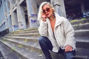 Фото тату: женщина в стильной одежде с татуировками на шее и груди, позирующая на фоне городской архитектуры.