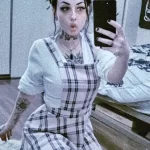 Девушка с татуировкой подсолнуха на предплечье, в клетчатой одежде и с ярким макияжем.