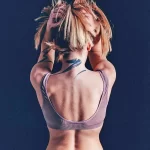 Фотография женщины с татуировкой в стиле акварель на спине и шее, изображающей абстрактные линии и мазки синего и черного цветов. Женщина держит волосы на голове.
