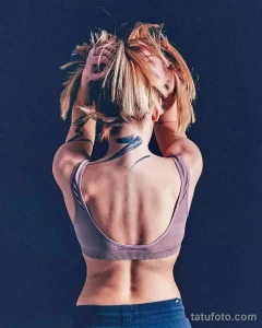 Фотография женщины с татуировкой в стиле акварель на спине и шее, изображающей абстрактные линии и мазки синего и черного цветов. Женщина держит волосы на голове.