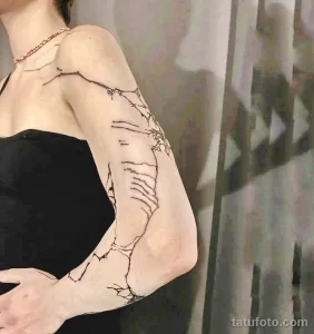Фото тату: изящный рисунок ветвей с цветами на плече. Эскиз татуировки выполнен в тонких черных линиях, создающих эффект акварели.