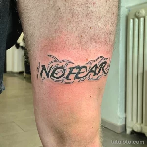 Фото тату: Надпись 'No Fear' на бедре, выполненная в готическом стиле с элементами пламени. Подходит для тех, кто хочет выразить свою смелость и решительность.