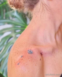 Небольшая татуировка в виде цветка с голубыми лепестками, расположенная на ключице девушки с веснушками. Изящный рисунок татуировки.